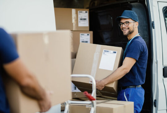 Young happy manual worker carrying cardboard boxes in delivery van while communicating with his colleagues.